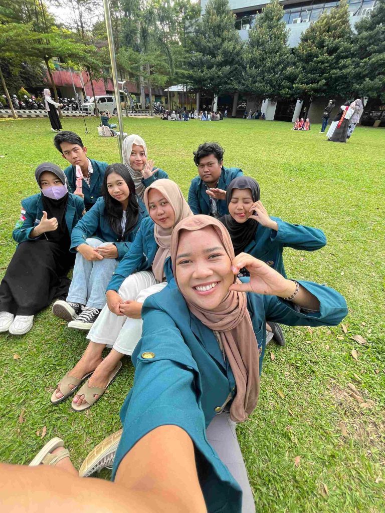 Mahasiswa KKN Kampung Baru UNILA duduk bersama di atas rumput, tersenyum dan berpose santai saat pengambilan gambar untuk reels perkenalan.