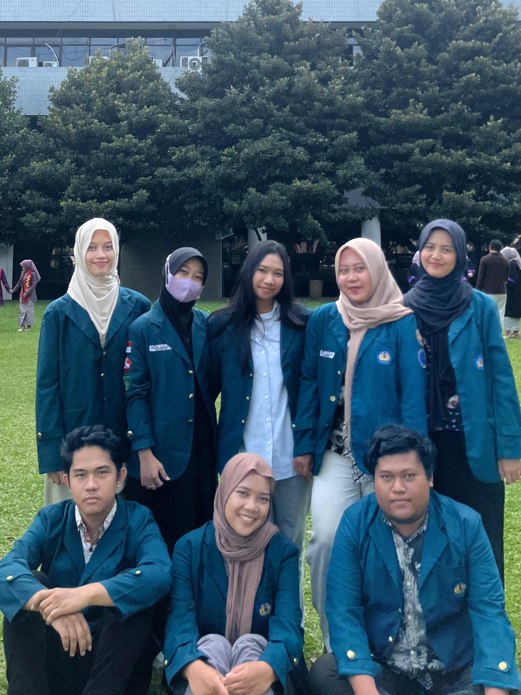 Sekelompok mahasiswa KKN Kampung Baru berfoto bersama di halaman belakang Gedung Rektorat dengan latar pepohonan dan bangunan kampus.